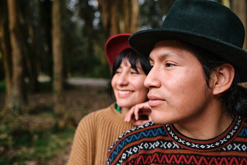 A Latin man wearing traditional clothes of his country and hat posing with his girlfriend behind him in the forest looking away.