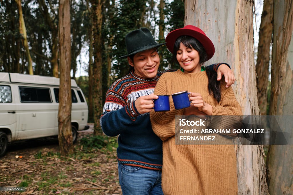 A happy couple toasting with metal mugs in the forest with the motorhome behind them A woman with her boyfriend enjoying a picnic with the motorhome behind having a hot drink in metal cups. Indigenous Peoples of the Americas Stock Photo