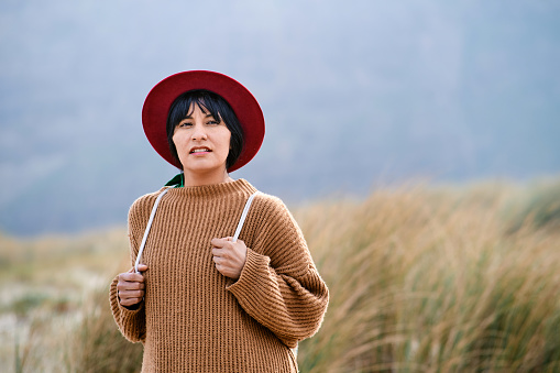 Front view of a Bolivian woman in red hat and sweater holding her backpack in tall grasses while looking away with copy space.