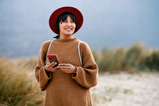 Front view of a smiling Latina woman in red hat holding her cell phone while looking away in nature.