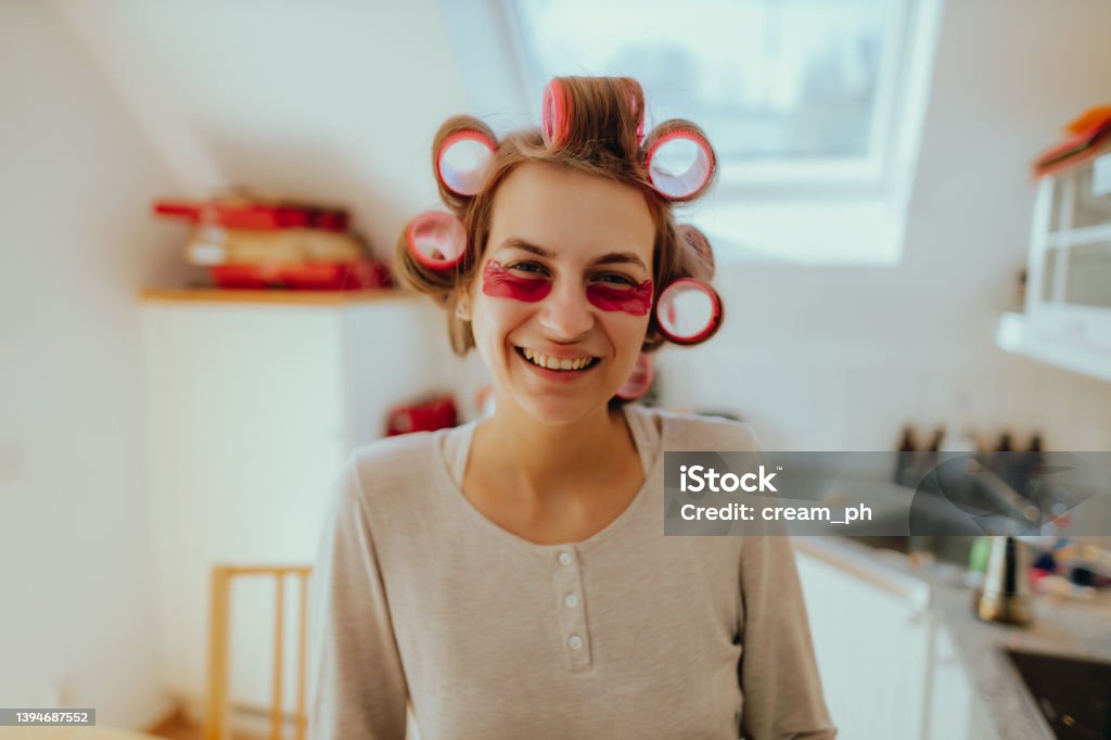 Smiling woman using hair rollers and an eye mask at home Portrait of a beautiful young smiling woman using hair rollers and an eye mask in the morning at home Facial Mask - Beauty Product Stock Photo