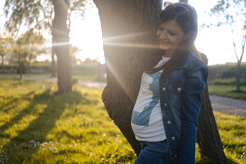 Beautiful pregnant woman stands in the park at sunset and hugs a tree. Merging with nature