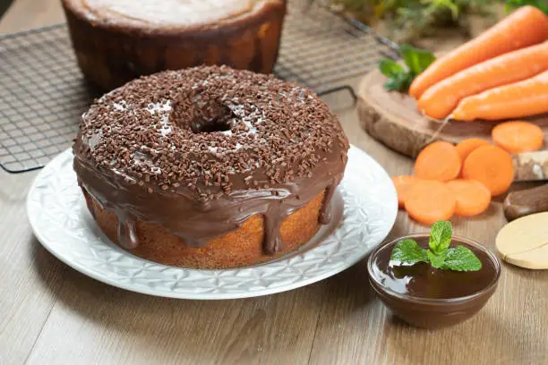 Brazilian carrot cake with chocolate frosting on wooden table with carrots in the background.