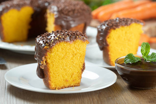 feche peça de bolo de cenoura brasileira com cobertura de chocolate em mesa de madeira com cenouras ao fundo - cake carrot carrot cake dessert - fotografias e filmes do acervo