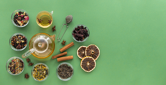 Several varieties of herbal and fruit tea in glass bowls and a kettle with a cup of hot tea on a green background. View from above. Healthy eating. Delicious warming drink. High quality photo