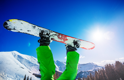 Snowboarder legs with board in the air upside down over Alpine tops and sunny sky on background