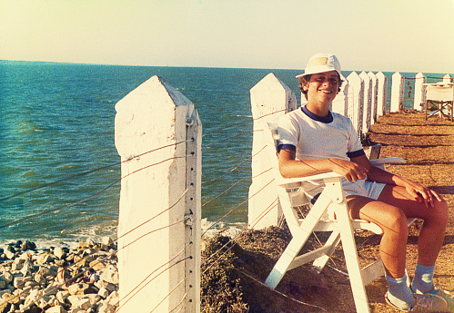 Analog photo of a teenage boy sitting by the sea. Summer vacation from the eighties. Grainy image from the eighties.