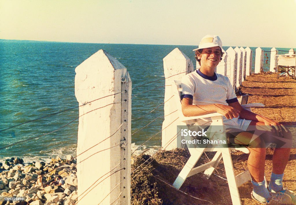 Vintage image of a teenager sitting by the sea Analog photo of a teenage boy sitting by the sea. Summer vacation from the eighties. Grainy image from the eighties. Retro Style Stock Photo