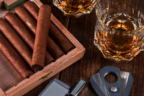 scatola con sigari cubani, accendino e taglierina sul vecchio piano del tavolo in legno. due bicchieri di whisky o alcol sullo sfondo. - cigar whisky bar cognac foto e immagini stock