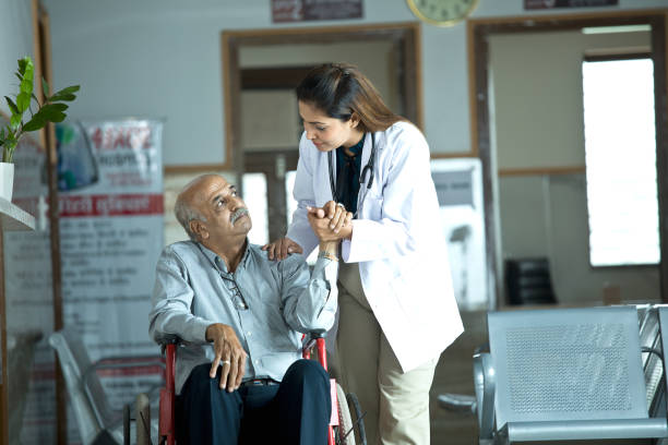 Female doctor consoling worried senior man sitting on wheelchair at hospital stock photo