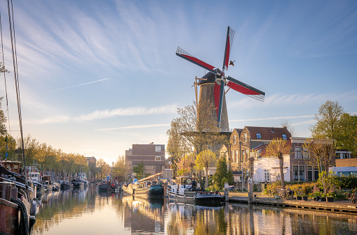 A harbor with boats reflecting in the water and a windmill towering over them. Beautiful sunlight and wispy clouds in the sky.