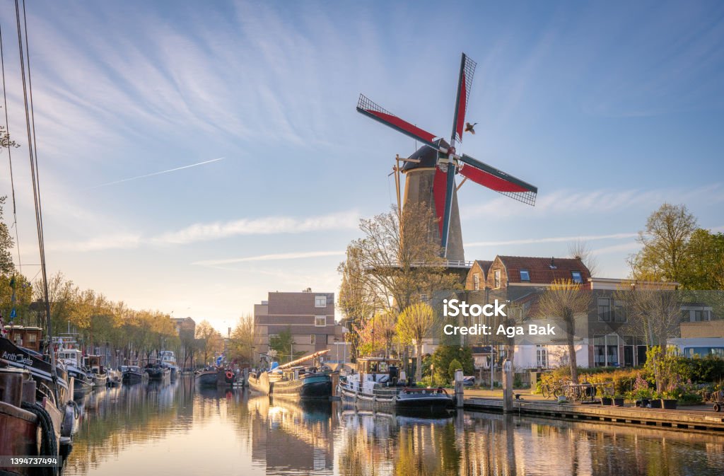 The golden hour in the city of Gouda, the Netherlands. A harbor with boats reflecting in the water and a windmill towering over them. Beautiful sunlight and wispy clouds in the sky. Gouda - South Holland Stock Photo