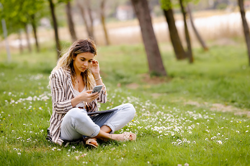 Young woman sitting on green grass, working on laptop and typing on mobile phone in city park on sunny spring day