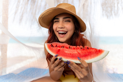 Pretty young girl in red bikini and straw hat in camellia on beach holding watermelon slice and smiling.
