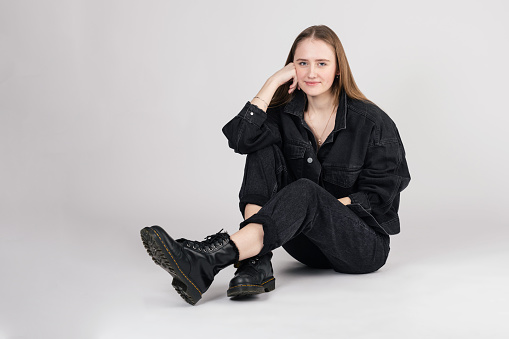 Fashion concept. Beautiful woman with long brown hair sitting on studio floor with one hand supporting the cheek in white background with copy space. Model wearing black jeans pants and jacket
