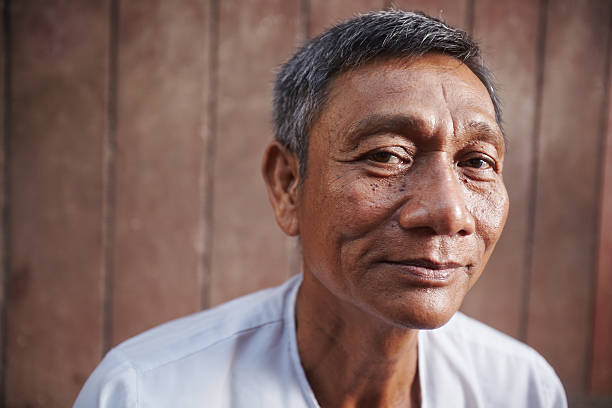 asian old man looking at camera against brown wall Portrait of happy mature asian man looking at camera against brown wall. Copy space cambodian ethnicity stock pictures, royalty-free photos & images