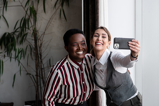 Two young businesswomen casually talking. Two young interracial coworkers smiling. Group of young mixed race colleagues on a smartphone. Interracial group of businesspeople taking a selfie.