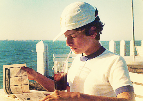 Analog photo of a teenage boy drinking a soft drink while reading outdoors by the sea during summer vacation from the eighties. Grainy image from the eighties.