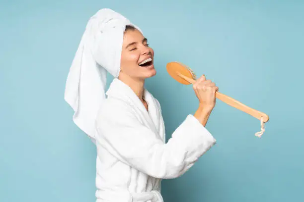 Photo of Young funny woman singing after shower in bathrobe and towel over head isolated on blue