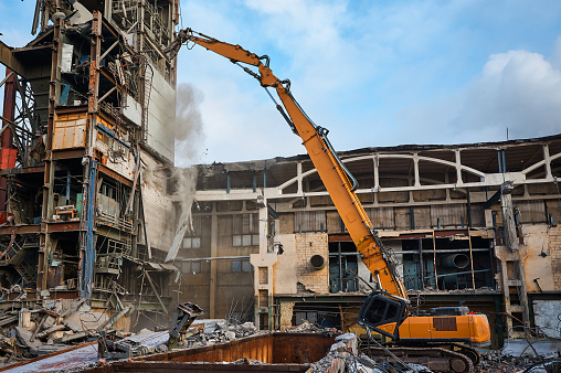 Destroying of old industrial building by modern high excavator destroyer at demolition site under blue sky on winter day