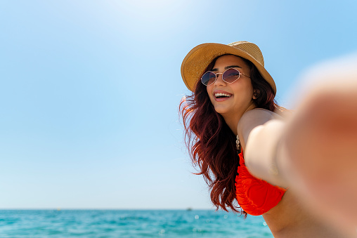 Smiling girl in straw hat and sunglasses take a selfie at the beach. Pretty young girl wearing red and yellow bikini sitting on the pebble beach and smiling.