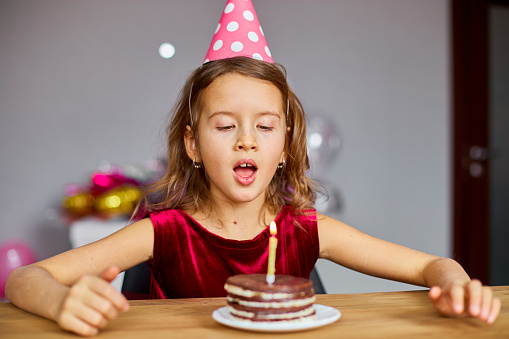 A little girl is wearing a birthday hat looking at a Birthday Cake and blow candles for a celebration party concept.