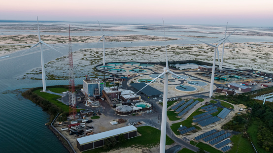 Sustainable energy is used in environmental conservancy. A wastewater treatment plant powered by wind turbines and solar panels near Atlantic City in New Jersey, USA. Aerial elevated view at the sunset.