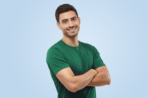 Portrait of attractive young sporty male in green t-shirt standing with crossed arms isolated on blue background. Fitness or gym trainer
