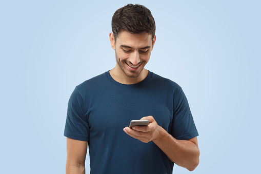 Indoor shot of good-looking young male looking at smartphone, smiling openly while holding smartphone in one hand