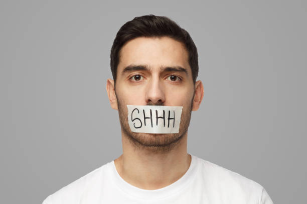 Young man with taped mouth with shhh text on it Close up shot of young man with taped mouth with shhh text on it finger on lips stock pictures, royalty-free photos & images