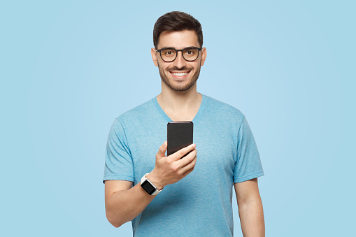 Young handsome man in blue t-shirt holding smartphone and looking at camera with smile while standing isolated on blue background