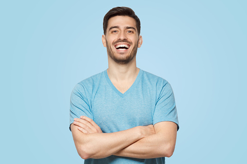Portrait of young positive guy wearing light blue T-shirt, standing with arms crossed, laughing happily at joke