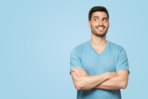 Portrait of young handsome guy wearing t-shirt standing with crossed arms, looking up and smiling with curious face, isolated on blue background with copy space