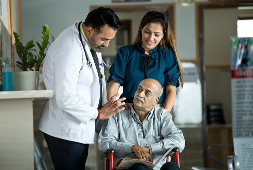 Male doctor and disabled old man in wheelchair with daughter by his side at the hospital corridor