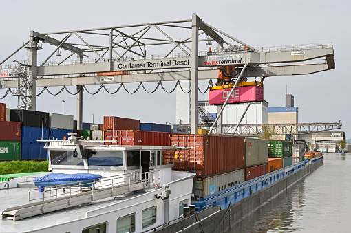 Basel, Switzerland - April 2022: Heavy crane in the city's container terminal unloading an industrial barge carrying shipping containers in the docks on the Rhine River