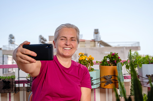 A 55-year-old woman with white hair and green eyes. He is sitting on the balcony of his house. Flowers you grow around her are in pots.Hobby joy of living model with positive emotion.Background is clear sky, space for text.Taken from a lower angle portrait model waist up.Model taken from front profile