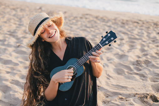 Beautiful young woman with long curly healthy hair wearing trendy straw hat playing ukulele at the beach.
