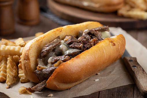 Philly cheesesteak sandwich made with steak, cheese and onions on a toasted hoagie roll with french fries on a wooden table