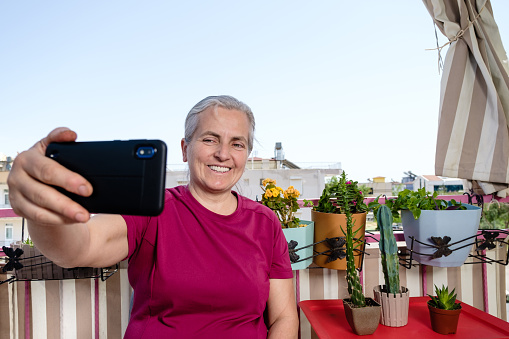 A 55-year-old woman with white hair and green eyes. He is sitting on the balcony of his house. Flowers you grow around her are in pots.Hobby joy of living model with positive emotion.Background is clear sky, space for text.Taken from a lower angle portrait model waist up.Model taken from front profile