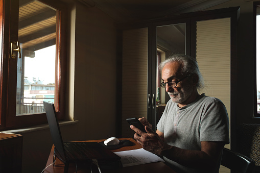 A man in his 60s, with long white hair, a slightly dirty white beard, and glasses. There is a computer on the table in the house. The model is sitting quite busy, taking notes with a phone in one hand and a pen in the other. The laptop is on. Sunlight enters through the window