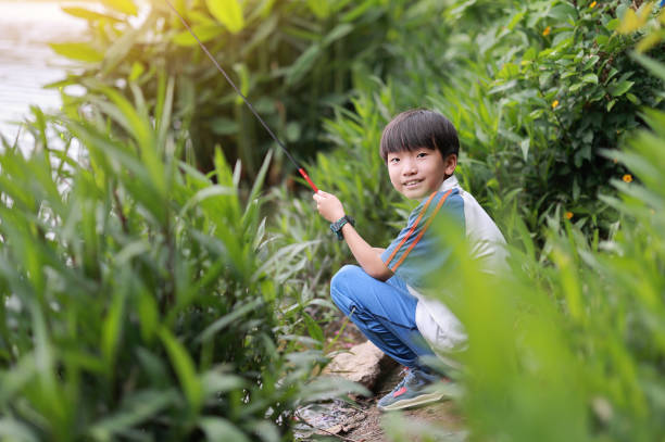 Child standing with fishing rod by calm river on summer day, enjoying outdoor family trip