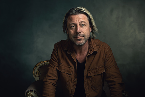 Man with blond hair and a stubble in a brown shirt sits on a sofa in front of a dark grey wall.