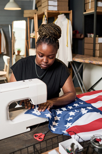 Young serious black woman in blue t-shirt sewing American flag while sitting in front of electric machine by large table in workshop
