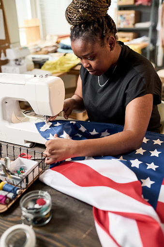 Young African American female volunteer making flag of USA in workshop while sitting by table with electric sewing machine