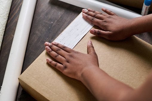 Hands of young African American female putting paper with client address on top of packed box with ordered goods