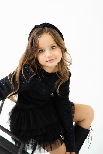 A little girl with blue eyes in a black jacket and black skirt is looking at the camera in the studio. Studio portrait