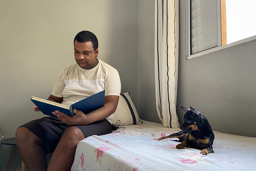 Portrait of a black young man at home, enjoying some leisure time. He is reading a book on his bed with the company of his little dog, a Pinscher.
