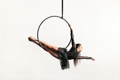 Full body of active barefoot female gymnast doing siren pose on hanging aerial hoop or lyra against white background in light studio