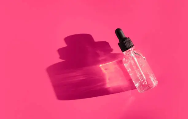 Transparent glass dropper bottle with air bubbles on bright pink purple background in the sunlight. Pipette with fluid hyaluronic acid, serum, retinol. Cosmetics, healthcare, beauty products concept