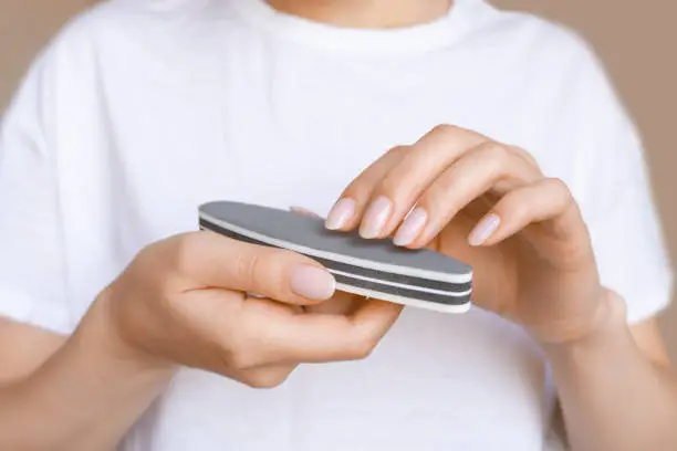 Female hands with natural manicure holding gray nail file. Woman in white holds emery board for smoothing and shaping the fingernails. Concept of self-care, beauty and professional tools usage